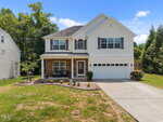 Charming two-story suburban house at 910 Woodland Road in Creedmoor with white siding, dark shutters, a covered front porch with seating, attached two-car garage, concrete driveway, grassy front yard, landscaped garden bed, and small bushes.