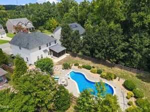 Aerial view of a two-story white house with a fenced backyard featuring a swimming pool, patio area with seating, landscaped bushes, and surrounding trees in a suburban neighborhood.
