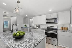 Modern kitchen with light gray cabinets, stainless steel oven and microwave, granite countertops, and a bowl of green apples on the island. Recessed lighting and a pendant fixture provide illumination.