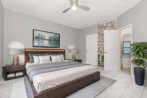 A modern bedroom with a large bed, two nightstands with lamps, a potted plant, a ceiling fan, and artwork above the headboard. The room has tiled floors, light gray walls, and two white doors.