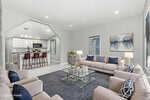 A modern living room with beige sofas, blue accent pillows, a glass coffee table, and a blue rug. The kitchen with a breakfast bar and pendant lights is visible through an open archway. Artwork hangs on the light gray wall.