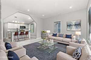 A modern living room with beige sofas, blue accent pillows, a glass coffee table, and a blue rug. The kitchen with a breakfast bar and pendant lights is visible through an open archway. Artwork hangs on the light gray wall.