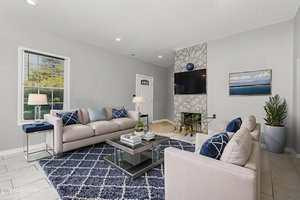 A modern living room with two beige sofas, a glass coffee table, a blue patterned rug, a wall-mounted TV above a fireplace, potted plants, and a window letting in natural light.