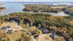 Aerial view of a residential area with large houses surrounded by trees, situated near a lake with several small peninsulas and wooded areas. The scene features winding roads and a mix of open grass and forested land.