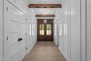 A hallway with light wood flooring, white paneled walls, and wooden ceiling beams leads to double front doors with glass panels. There are multiple white doors with black handles on the left wall.