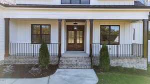 A front view of a house with a covered porch, double wooden doors, black framed windows, brick steps, metal railings, and landscaped shrubs in front of the porch.
