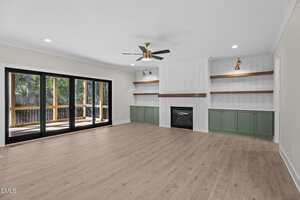 A spacious living room with light wood flooring, a ceiling fan, built-in shelves with green cabinets, a fireplace, and large sliding glass doors opening to a wooden deck with a view of trees.