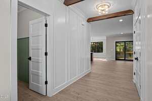 A hallway with light wood flooring, white paneled walls, and visible wooden ceiling beams leads to a living area with large windows and glass doors that open to a deck. A small green accent wall is visible in an adjoining room.