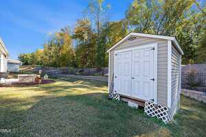 A light-colored storage shed with double doors and a gable roof stands on a grassy lawn near a house, with trees and a stone retaining wall in the background on a clear day.