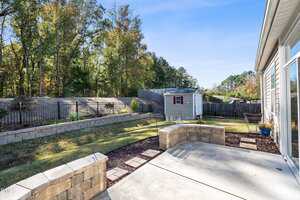 A backyard with a concrete patio, stone retaining walls, a landscaped lawn, a small gray shed, and a black metal fence. Trees and a clear blue sky are visible in the background.