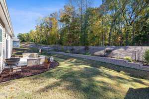 A backyard with a small circular patio featuring two chairs and a fire pit, bordered by mulch and grass. A retaining wall and black metal fence separate the yard from a wooded area beyond.