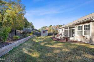 A backyard with green grass bordered by a retaining wall and landscaped plants, featuring a small shed, black metal fence, and a beige house with large windows and a screened patio on the right.