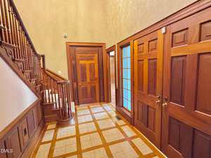 A foyer with wooden paneling, two closed wooden doors, a glass-paned door, and a staircase with wooden railings. The floor features a checkered tile pattern with wood trim. Walls are painted in a light neutral tone.