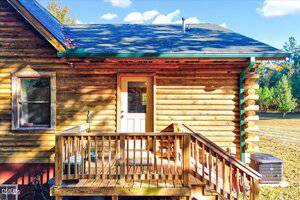A small wooden deck with stairs leads to a white door on the side of a log cabin. The cabin has a shingled roof, gutters, and sits in a yard with green grass and trees in the background.