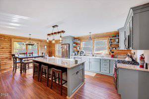 A spacious kitchen with light blue cabinets, a large island with three stools, stainless steel appliances, wood floors, and log walls. A dining table sits by a window, and two pendant lights hang from the ceiling.