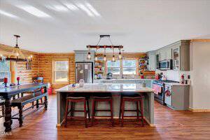 A spacious kitchen features a large island with three stools, gray cabinets, stainless steel appliances, wood floors, open shelving, and a dining area with a dark table and benches. Log walls and large windows are visible.