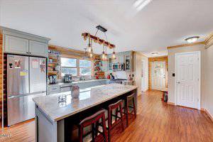 Modern kitchen with wood floors, light gray cabinets, a large marble island with three stools, pendant lights, stainless steel appliances, a window over the sink, and a door leading outside. Shelves display mugs and kitchen items.