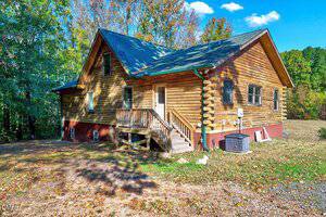A wooden log cabin with a green roof sits on a concrete foundation at 8545 Grassy Creek Road, Oxford. Front steps lead to a white door, with an HVAC unit outside and trees with autumn leaves in the background under a blue sky.