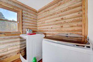 A laundry room with wood-paneled walls, a front-loading washer and a top-loading dryer. Laundry supplies are on top of the washer, and a bottle is on the floor. Sunlight comes in through a window on the left.