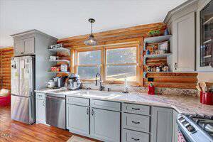 A kitchen with light gray cabinets, stainless steel appliances, a double sink under three large windows, open wood shelves holding dishes and appliances, and a log wall backdrop. Sunlight streams in through the windows.