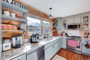 A kitchen with light gray cabinets, wood-paneled walls, and marble countertops. Appliances include a coffee maker, air fryer, gas stove, and microwave. Sunlight comes in through three large windows above the sink.