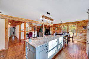 A spacious log cabin kitchen with wood floors, a large marble island, pendant lights, and dining area. Stairs and a workspace are visible in the background, with large windows letting in natural light.