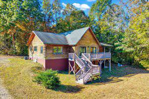 A wooden cabin with a raised front porch and staircase sits surrounded by trees with green and autumn foliage under a bright blue sky with scattered clouds.