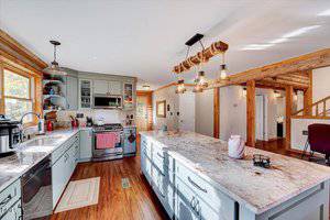 A spacious kitchen with a large marble-topped island, gray cabinets, stainless steel appliances, wood flooring, and rustic wood beams on the ceiling. Pendant lights hang above the island.