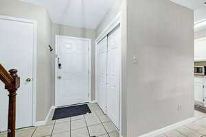 The image shows a tiled entryway with a white front door, two white closet doors, a small black doormat, beige walls, and a partial view of a staircase and kitchen area. Light switch and hook are visible on the walls.