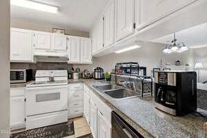 A kitchen with white cabinets, granite countertops, a white stove and oven, microwave, stainless steel sink, coffee maker, and various small appliances. Decorative items and utensils are placed on the counters and shelves.