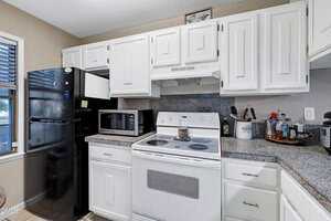 A kitchen with white cabinets, a black refrigerator, a white electric stove with oven, a microwave, granite countertops, and various kitchen items on the counters. A window is visible to the left.