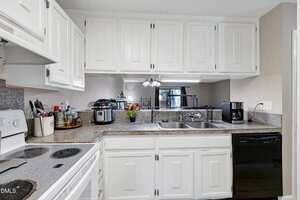 A kitchen with white cabinets, granite countertops, and stainless steel double sink. Appliances include a stove, microwave, slow cooker, toaster, and dishwasher. Small plants and kitchen items are on the counter.