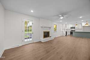Open-plan living area with light wood flooring, white walls, a brick fireplace, ceiling fan, recessed lighting, and a door leading outside. The kitchen features white cabinets, stainless steel appliances, and pendant lights.
