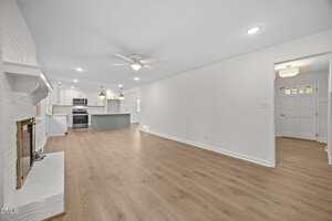 Open-concept living room with light wood flooring, white walls, ceiling fans, recessed lighting, a white brick fireplace, and a view into a modern kitchen with an island and stainless steel appliances. Entryway visible on the right.