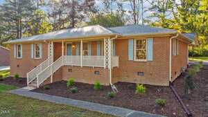 A single-story brick house at 102 Royall Road, Oxford, features light blue shutters, a covered front porch with white railings and stairs. The landscaped yard has mulch, small plants, and tall trees in the background.