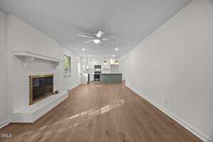 A spacious open-concept living area with light wood floors, white walls, a white brick fireplace on the left, recessed lighting, ceiling fan, and a modern kitchen with island and pendant lights in the background.