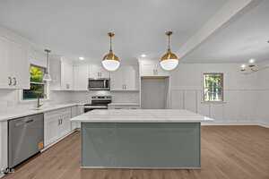Modern kitchen with white cabinetry, stainless steel appliances, light wood flooring, a large island with a gray base and white countertop, two gold pendant lights, and windows letting in natural light.