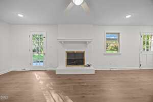 A bright living room with light wood floors, a white brick fireplace with a brass-trimmed glass door, a ceiling fan, and large windows and doors allowing natural light to enter.