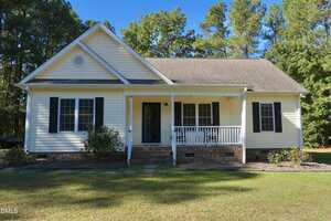 A single-story house with cream-colored siding, black shutters, a covered front porch with white railings, and a brick foundation, set in a grassy yard with trees in the background.