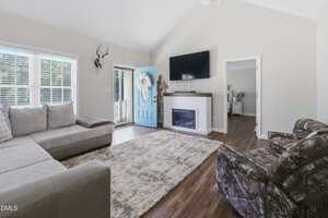 A living room with a gray sectional sofa, a large area rug, a mounted TV above a white fireplace, a camouflage armchair, and an open blue front door. Sunlight enters through large windows on the left.