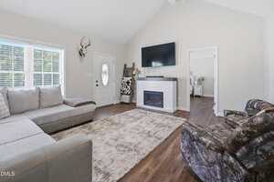 A living room with a sectional sofa, patterned recliner, wall-mounted TV above a fireplace, a beige rug on wood flooring, antlers on the wall, and a door with a circular window next to two large windows.