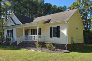 A single-story yellow house with white trim, a covered front porch, black shutters, and a brick foundation, surrounded by a lawn and trees under a clear blue sky.