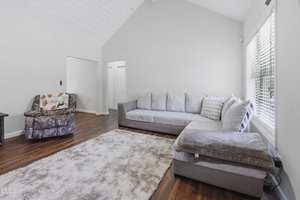 A living room with a large light gray sectional sofa, a small floral-patterned armchair with a blanket, a soft beige area rug, hardwood floors, white walls, and a large window letting in natural light.