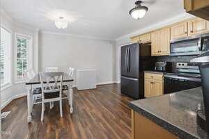 A kitchen with wood cabinets, black appliances, and dark countertops. A dining table with four chairs is near a window. There is a white chest freezer against the wall, and the floor is dark wood.