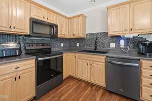 A modern kitchen with light wood cabinets, black appliances including a stove, microwave, and dishwasher, a black tile backsplash, countertop appliances, and a dark sink, all set on a wood floor.