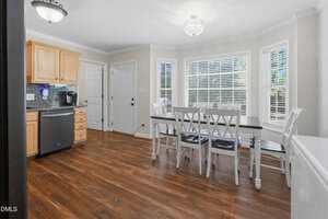 A kitchen and dining area with wood floors, light-colored walls, a dining table with six chairs, a dishwasher, light wood cabinets, and multiple windows with blinds allowing natural light into the room.