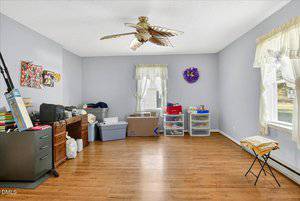 A spacious room with light purple walls, two windows, wooden floor, ceiling fan, and miscellaneous items including boxes, bins, a desk, and plastic drawers arranged along the walls.