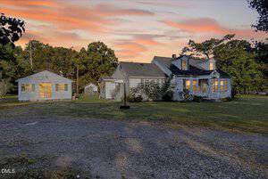 A single-story gray house with a pitched roof and white trim sits on a grassy lot at sunset at 8663 US Hwy 15 N in Bullock. A detached smaller building is nearby, with a gravel driveway and trees in the background.