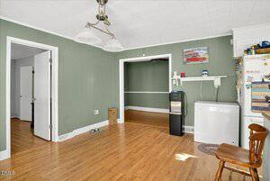 A room with green walls and wood flooring features a chest freezer, water dispenser, small wall shelf, white fridge, and wooden chair. Two open doorways lead to other rooms. Pet dishes and a cardboard box are on the floor.