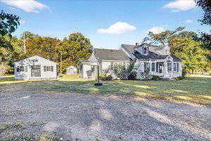 A light gray house with white trim and a detached matching building sit on a large grassy lot with trees in the background and a gravel driveway in the foreground under a blue sky with a few clouds.